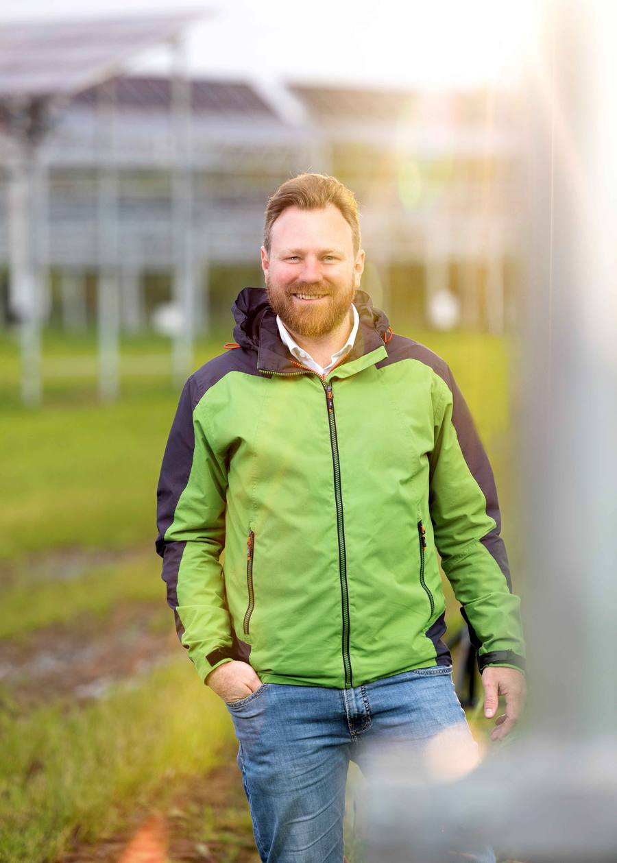 Lächelnder Mann in grüner Jacke zwischen Modulreihen der Agri-PV-Anlage in Jülich