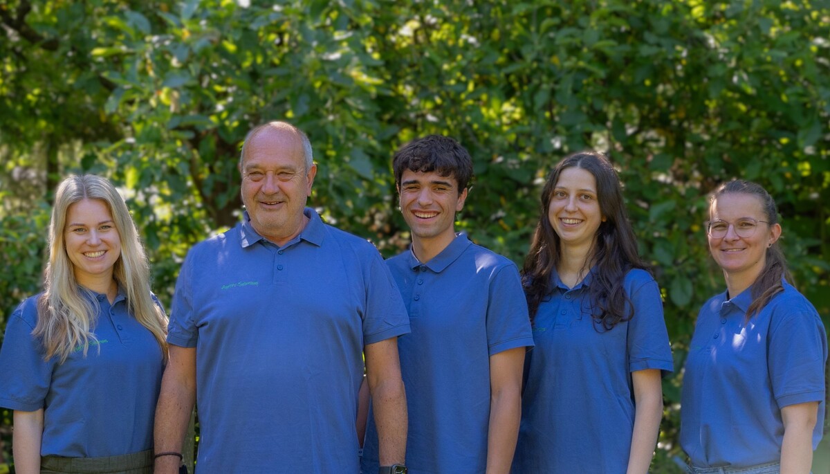 Fünfköpfiges Team in blauen Poloshirts lächelt gemeinsam vor grünem Blattwerk im Freien.