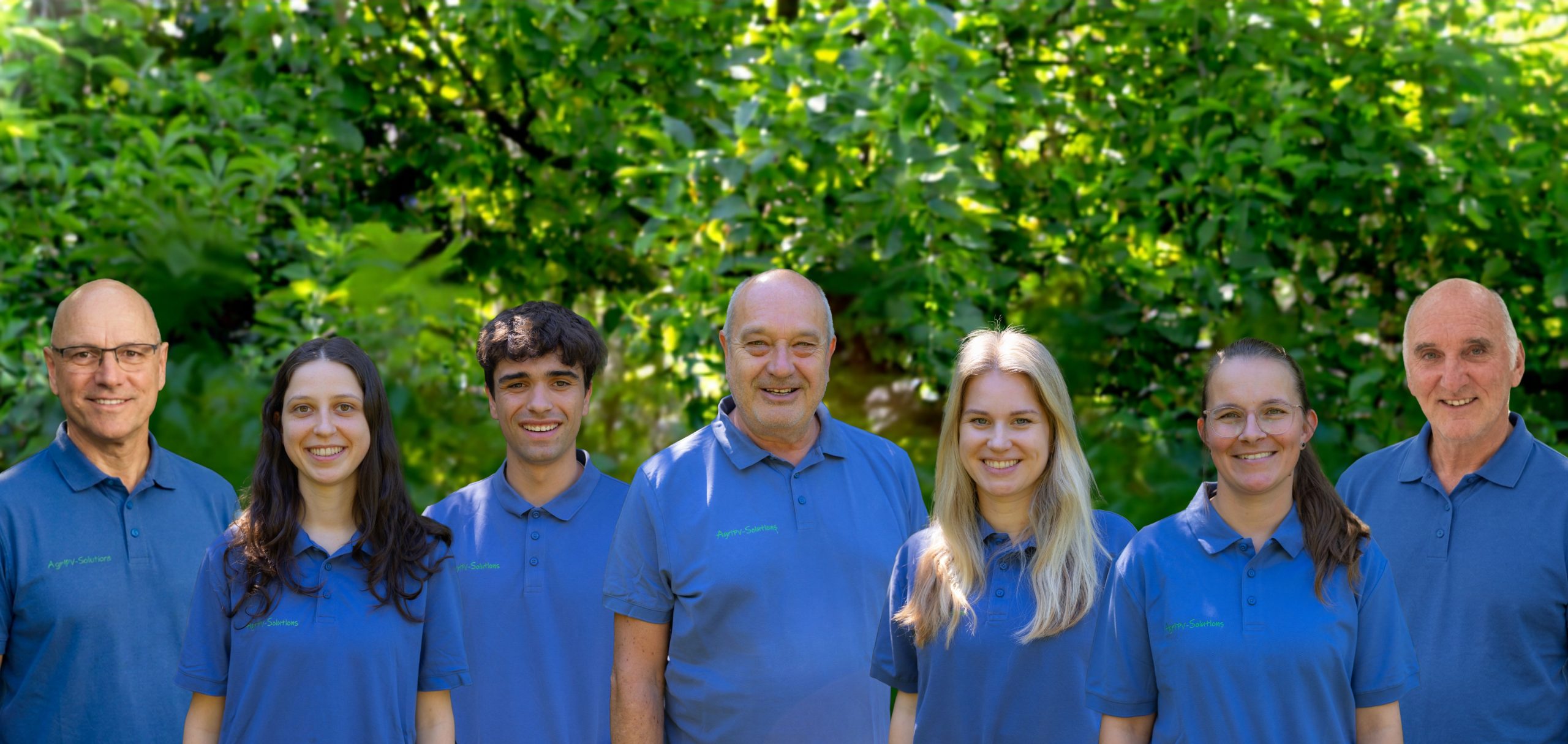 Fünfköpfiges Team in blauen Poloshirts lächelt gemeinsam vor grünem Blattwerk im Freien.